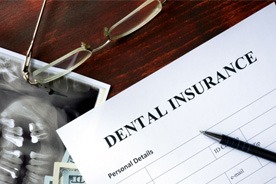 A dental insurance form sitting on a wooden desk next to a pen, glasses, and X-rays